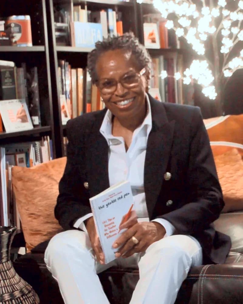 Bibi Angola in a navy blazer holding The Gentle Red Pen, her Teacher of the Year poster behind her