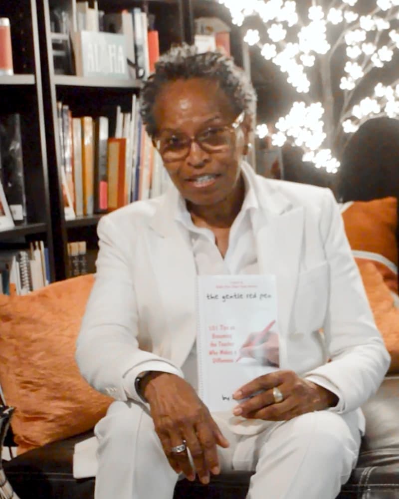 Bibi Angola in a white suit, smiling and holding The Gentle Red Pen