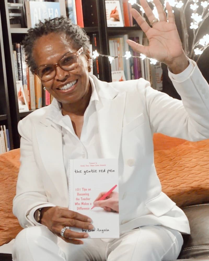 Bibi Angola in a white suit, smiling and waving, holding The Gentle Red Pen